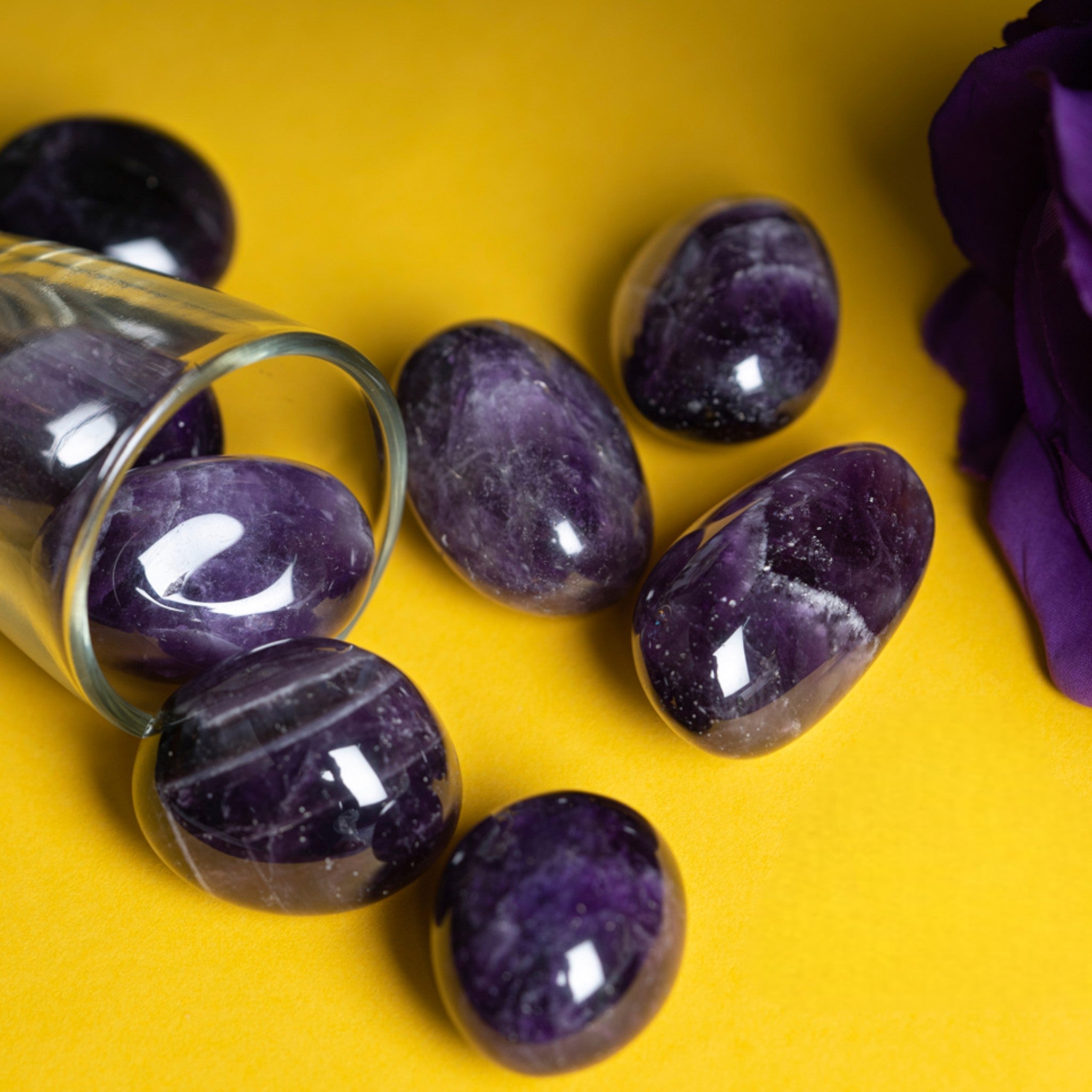  polished amethyst stones spilling out of a glass jar onto a yellow surface, with a purple fabric to the side.