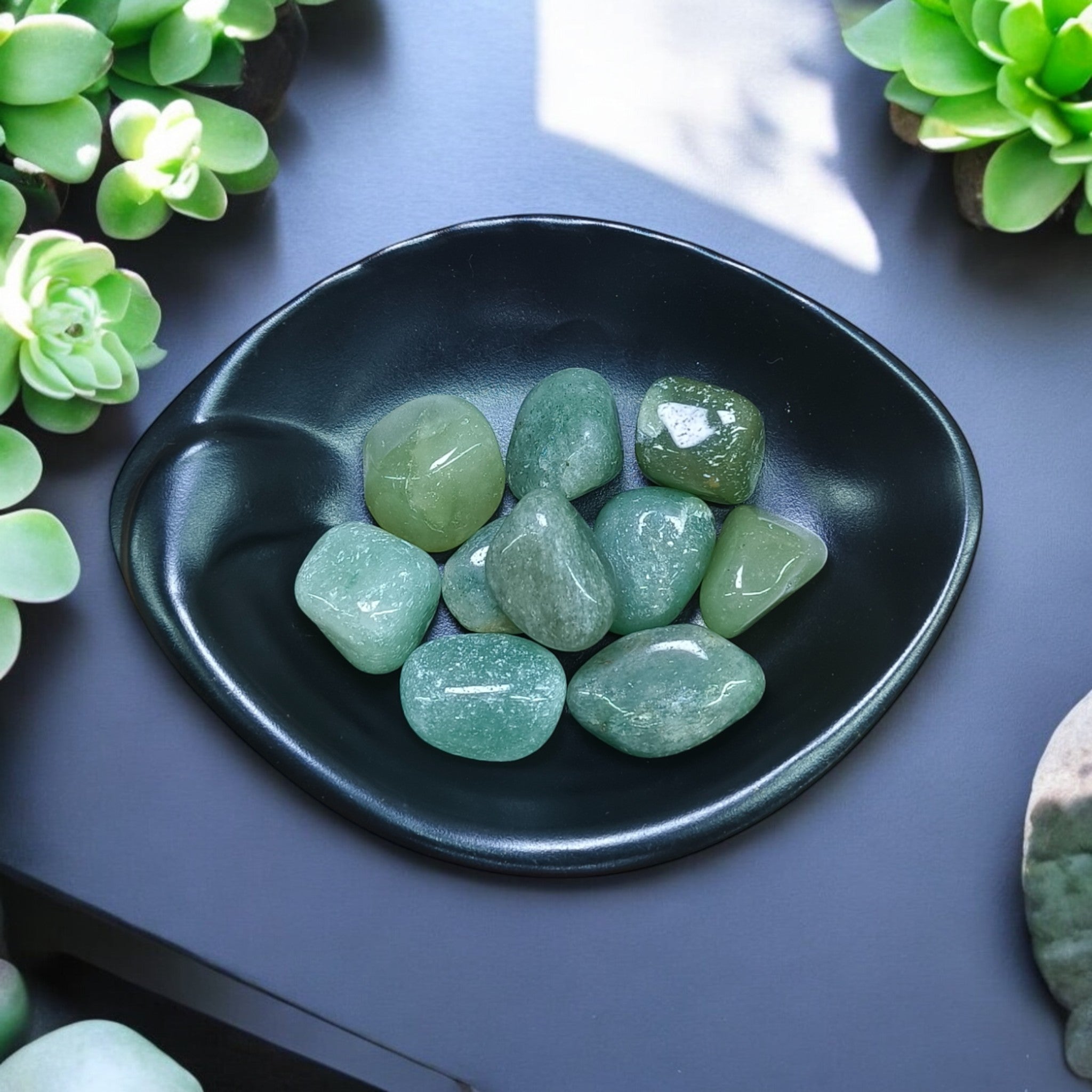 A collection of polished green aventurine stones arranged on a black plate, with succulent plants in the background.