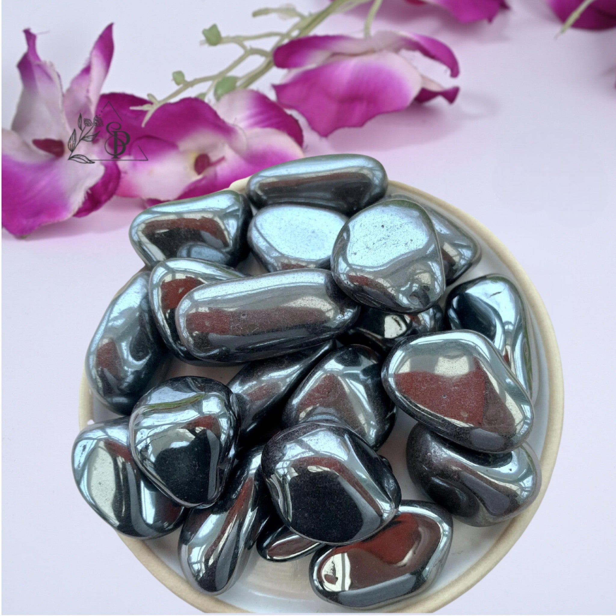 A collection of polished hematite stones displayed in a white bowl, with a pink flower in the background.
