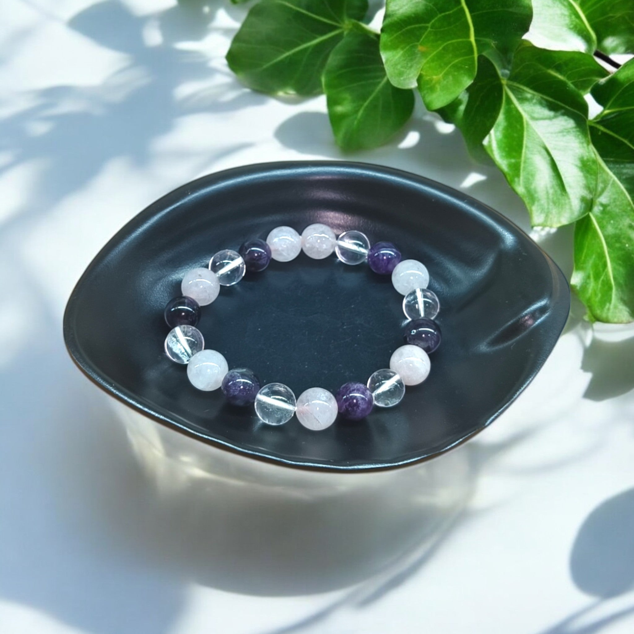 “Rose Quartz, Clear Quartz and Amethyst bracelet in 10 mm natural beads – pastel crystal trio bracelet in black dish and green leaves in background"