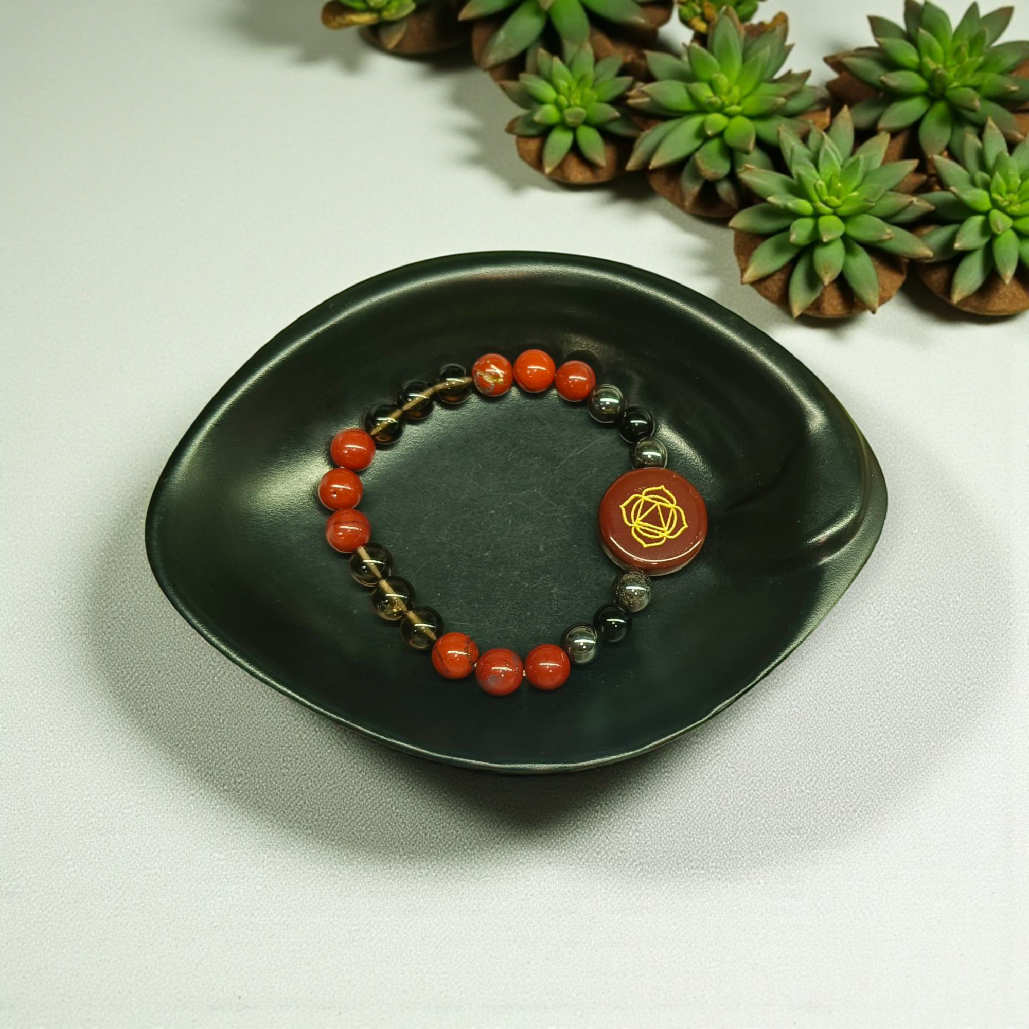"Root Chakra bracelet with engraved Red Jasper round dial centerpiece and crystal beads of Red Jasper, Smoky Quartz, Hematite, and Black Obsidian in black bowl"