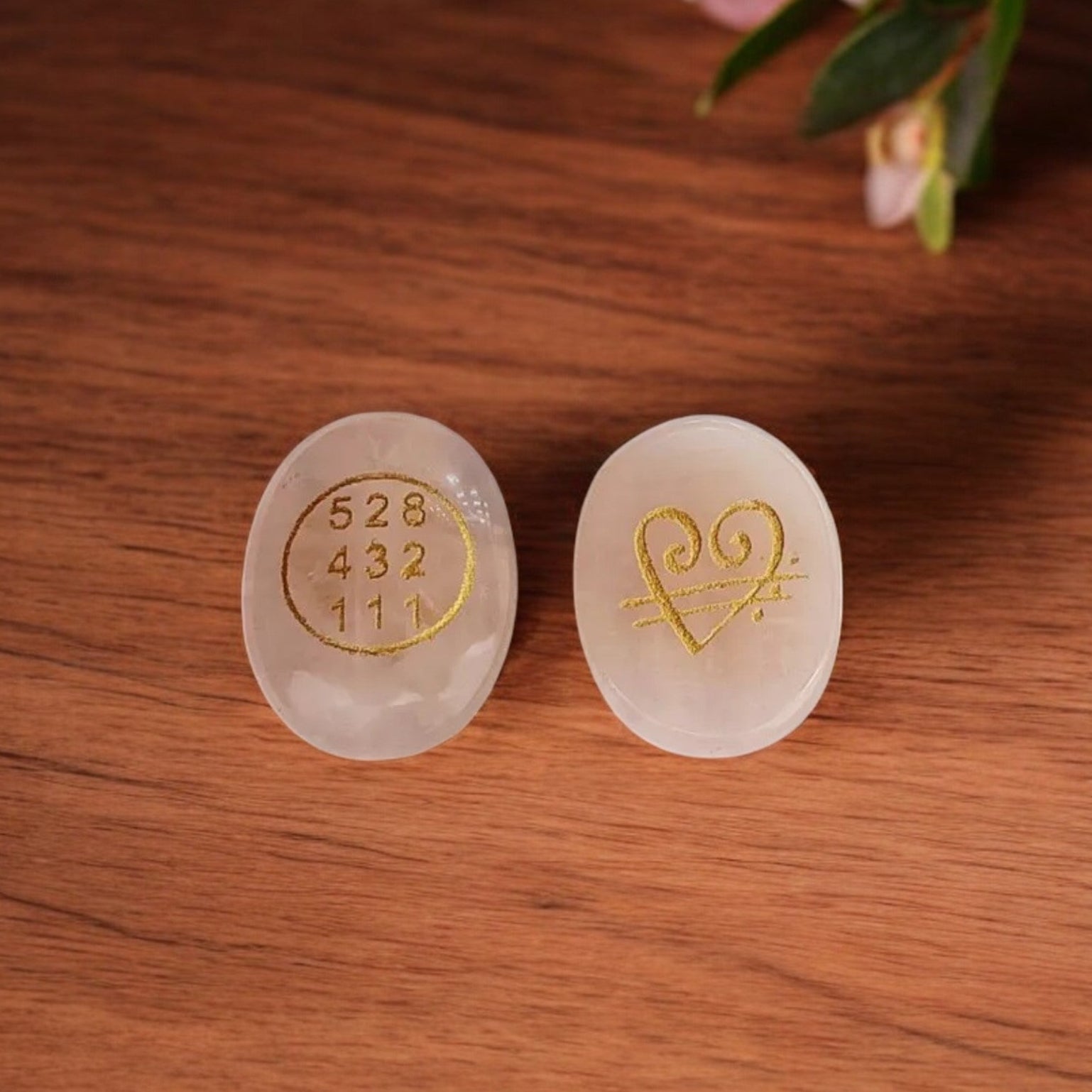 Rose quartz stones with golden engravings of number and heart symbol showing front and back of the same stone on a wooden surface with flowers in the background