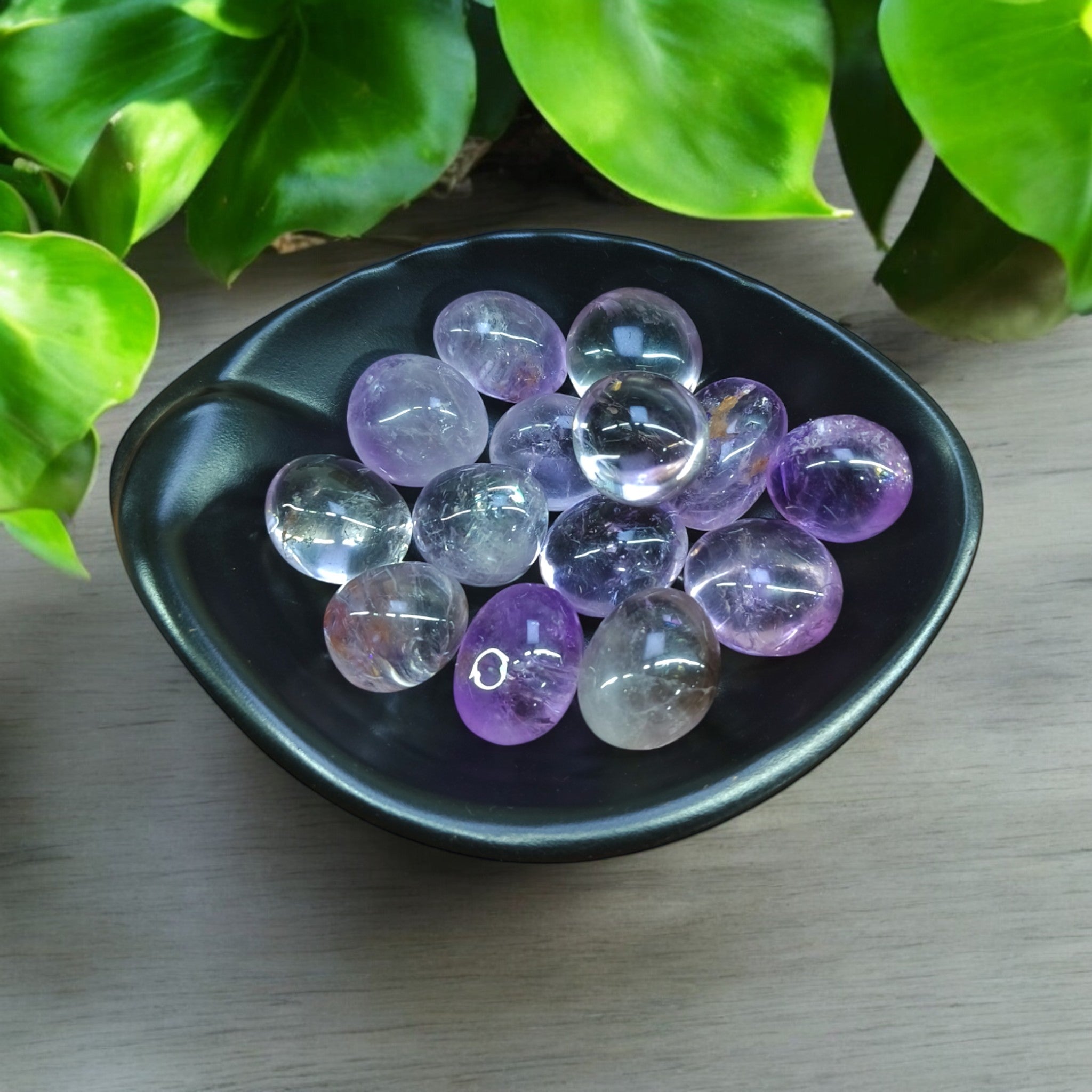 A collection of polished amethyst stones arranged in a black bowl, with a few leaves in the background providing a natural feel.